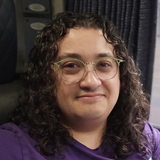 photo of TTP, a white person with long, curly brown hair, a round face, and stylish green rimmed glasses. They are smiling and seem deeply content.