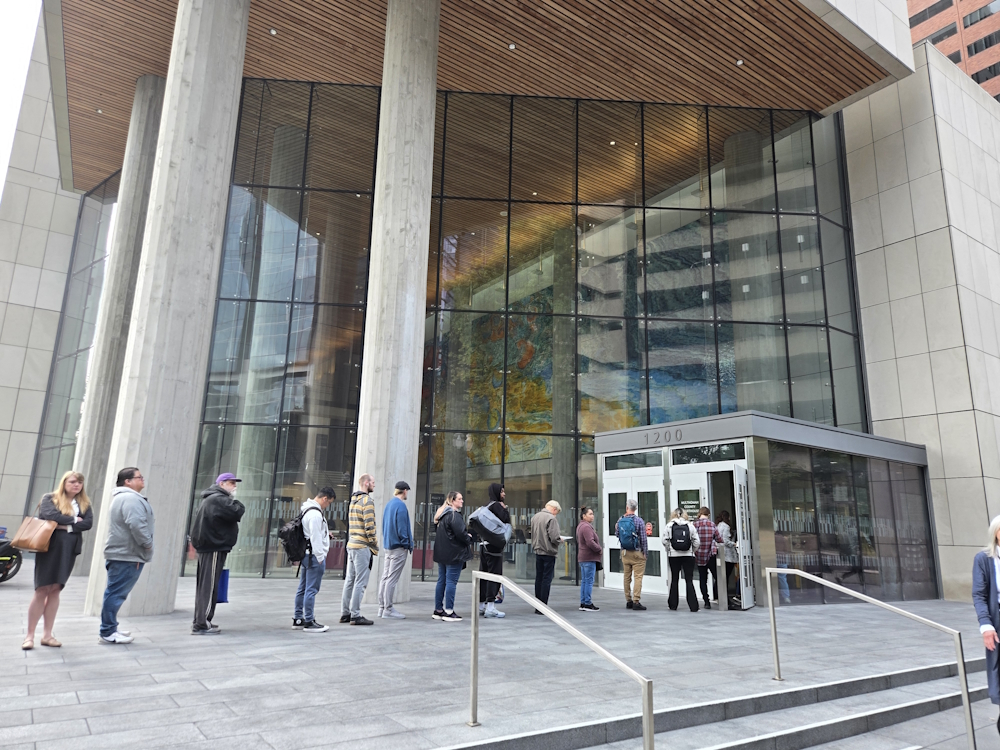 People standing in line to enter the Multnomah County Courthouse. Two imposing concrete columns and an enormous glass facade loom over the people.